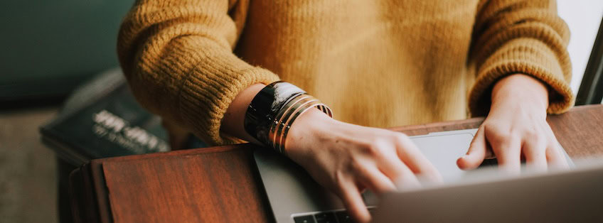 Person in einem gelben Wollpullover arbeitet an einem Laptop, der auf einem Schreibtisch aus dunklem Holz steht. Foto von Christian Hume von Unsplash.